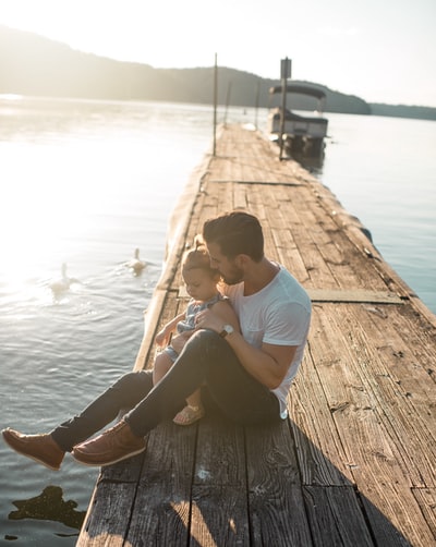 白天，男人和女孩坐在船和两只白鸭子附近的棕色码头上